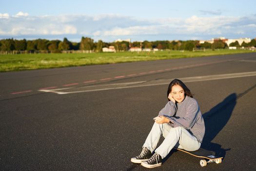 Skater girl sits on her skateboard on road, using smartphone, chatting on mobile app
