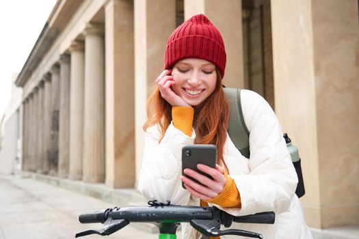 Redhead girl, tourist with backpack, uses mobile phone to rent e-scooter on streets of European city