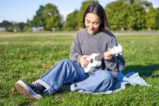 Carefree asian girl singing and playing ukulele in park, sitting on grass, musician relaxing on her free time outdoors