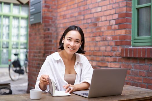 Working woman sitting in coworking space, drinking coffee and using laptop, wearing wireless headphones, watching video and smiling