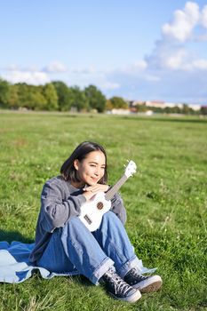 Romantic asian girl sitting with ukulele guitar in park and smiling, relaxing after university, enjoying day off on fresh air