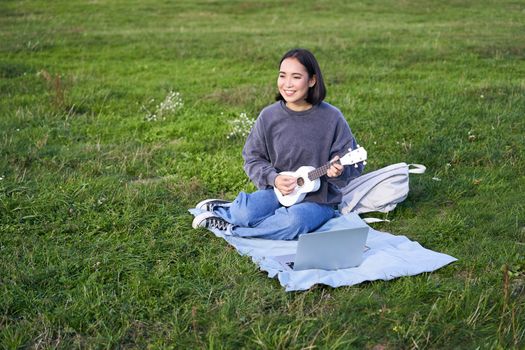 Cute korean girl, musician sits in park, plays ukulele and sings, looks up chords and tutorials on laptop