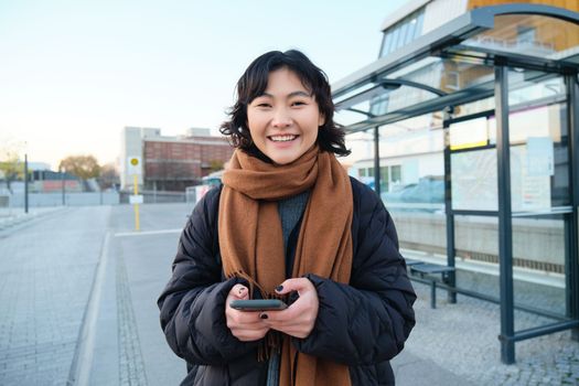 Portrait of Korean woman in winter jacket, standing with smartphone, waiting for bus on stop, looking at mobile phone app checking public transport application.