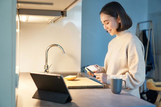 Portrait of young korean woman, making toast and watching video on digital tablet, looking at screen