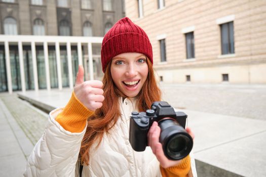 Beautiful readhead girl, photographer with professional camera takes pictures outdoors, walking around city and taking photos, sightseeing