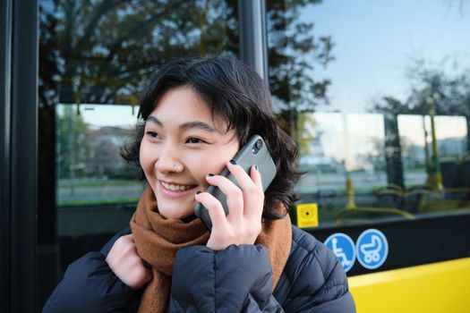 Cellular technology and people concept. Stylish asian girl talks on mobile phone, makes a telephone call, stands near bus stop and has conversation