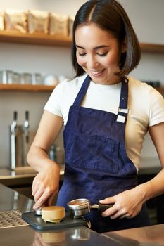 Vertical shot of smiling asian girl putting coffee in tamper to brew capuccino, prepare order in coffee machine, standing behind counter in blue apron