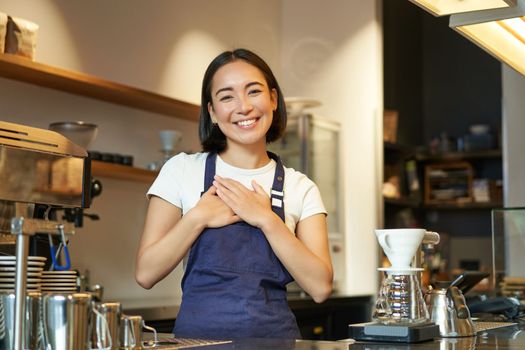 Friendly smiling asian girl barista, holds hands on heart, looks grateful at client, loves her guests in cafe, stands in uniform apron