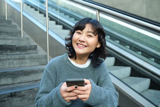 Portrait of brunette young woman, student sits on stairs in public place, looks at smartphone, reads text message and smiles
