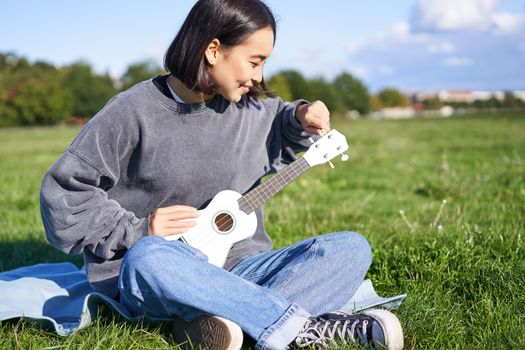 Singing asian girl playing ukulele on grass, sitting on blanket in park, relaxing outdoors on sunny day