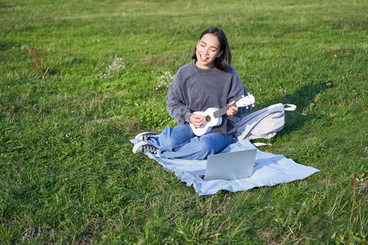 Cute korean girl, musician sits in park, plays ukulele and sings, looks up chords and tutorials on laptop