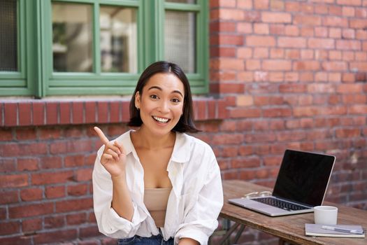 Asian girl with surprised face, sitting with laptop, pointing left at copy space, banner with information, looking amazed