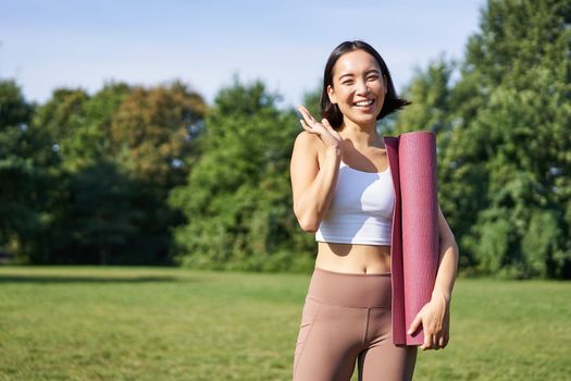 Happy asian fitness girl says hi, waves hand, stands with rubber yoga mat for workout in park, wears sport uniform.