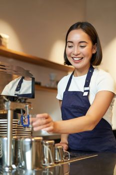 Portrait of cute barista girl working behind counter, making coffee, steaming milk for cappuccino, wearing cafe uniform blue apron