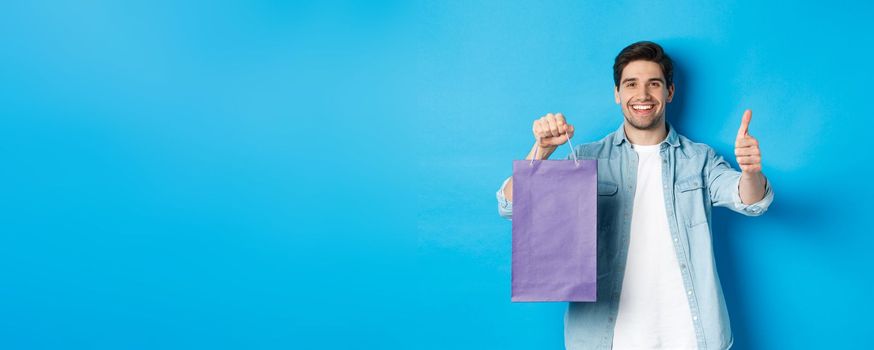 Concept of shopping, holidays and lifestyle. Satisfied smiling man holding paper bag, showing thumb-up and recommending store, standing over blue background
