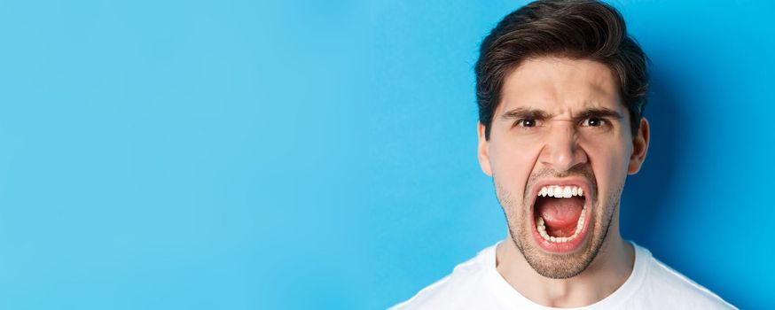 Head shot of angry man shouting and looking with hatred, standing mad against blue background
