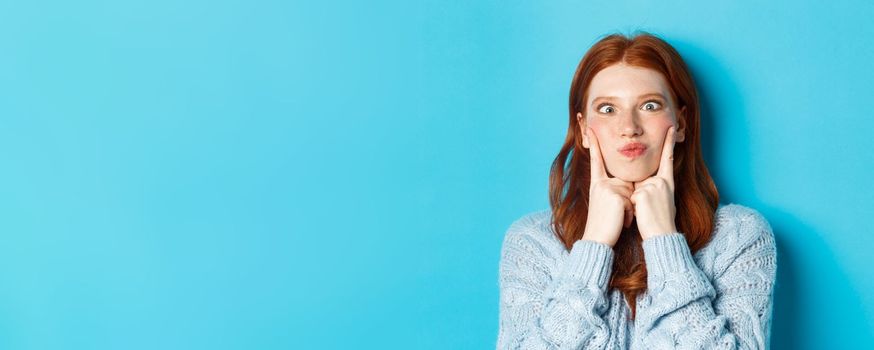 Close-up of funny redhead teen girl making faces, squinting and pocking cheeks, standing against blue background