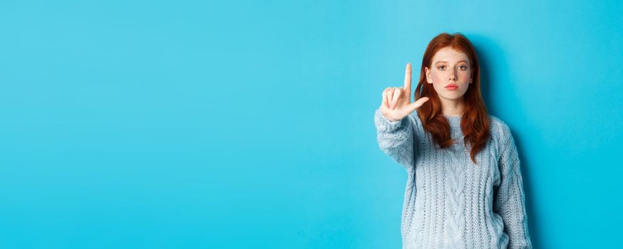 Serious redhead girl in sweater showing taboo gesture, extending one finger, shaking forefinger to disapprove, disagree and forbid something, blue background