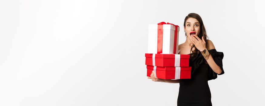 Celebration and christmas holidays concept. Woman holding xmas gifts and looking surprised, receive presents, standing over white background