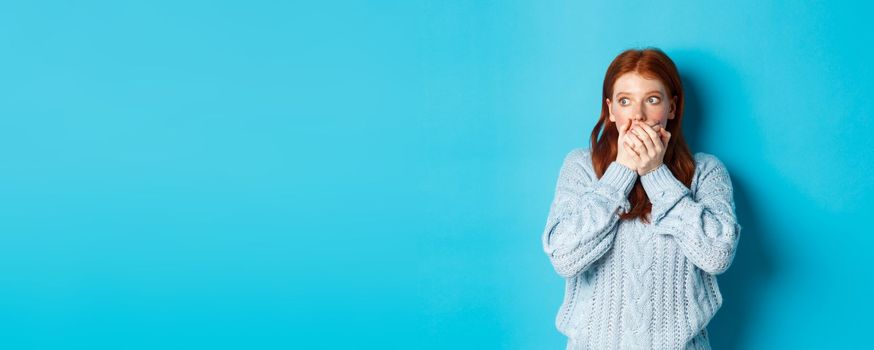 Shocked and anxious redhead girl staring left scared, covering mouth and gasping, standing over blue background in sweater