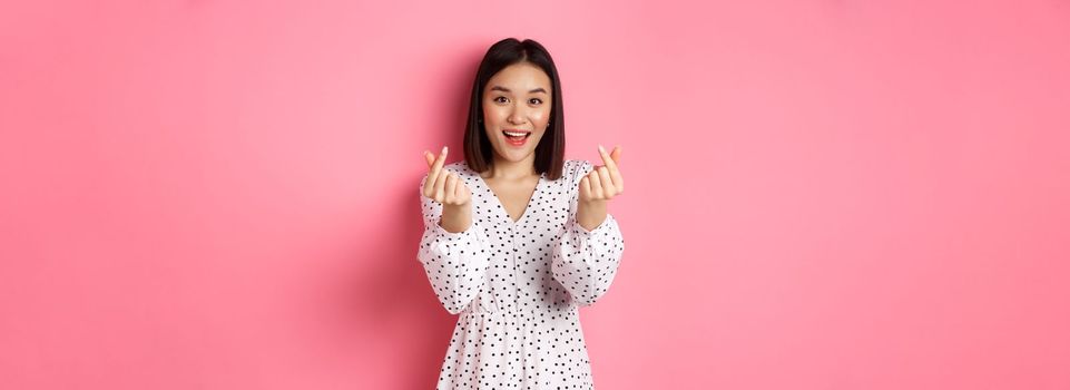Lovely asian woman in dress showing korean heart signs and smiling, standing on romantic pink background