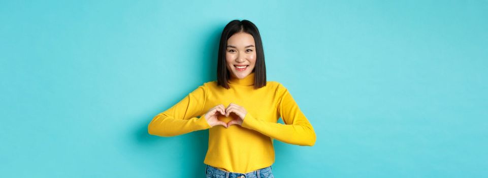 Valentines day and romance concept. Beautiful asian woman show I love you, heart gesture and smiling, standing against blue background