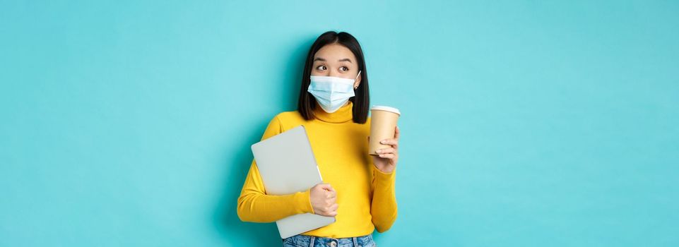 covid-19, health care and quarantine concept. Asian girl student in medical mask standing with laptop and coffee from cafe, standing over blue background