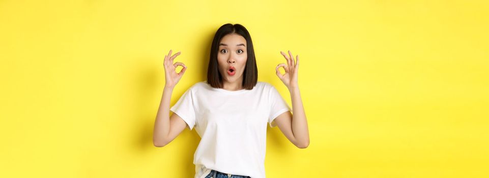 Impressed korean girl saying WOW, showing okay signs and looking amazed, standing against yellow background