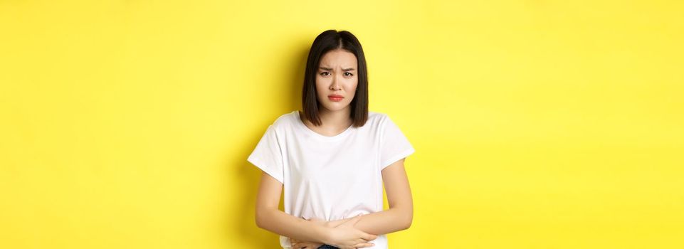 Image of asian woman feeling sick, having painful cramps, holding hands on belly and frowning from pain, discomfort during menstrual period, standing over yellow background