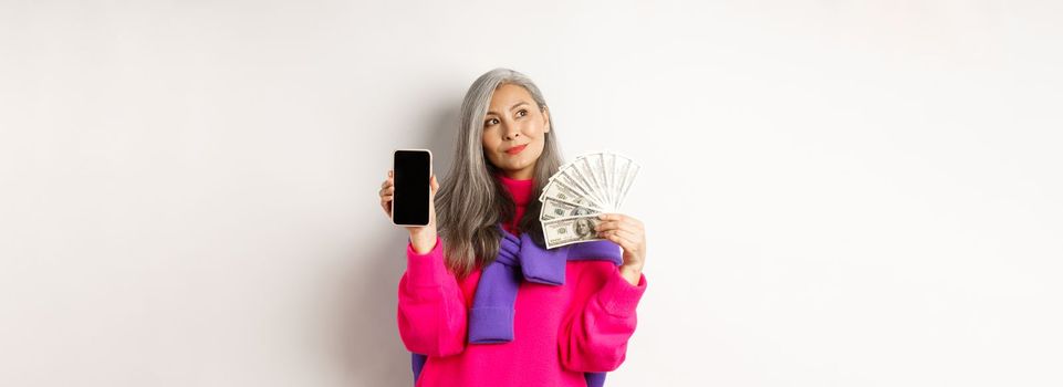 Fashionable asian senior woman showing money dollars and blank smartphone screen, demonstrate online shop, standing over white background