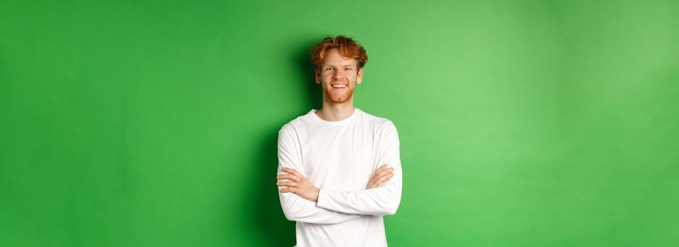 Young ambitious man with red hair standing over green background, holding hands crossed on chest and smiling