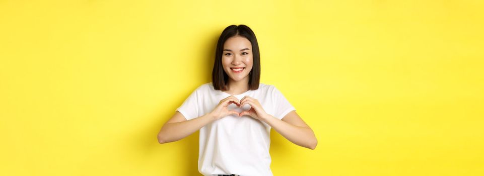 Beautiful asian woman showing I love you heart gesture, smiling at camera, standing against yellow background. Concept of valentines day and romance