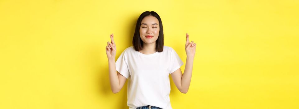 Hopeful asian girl making wish, cross fingers for good luck and praying with eyes closed, saying please, standing over yellow background