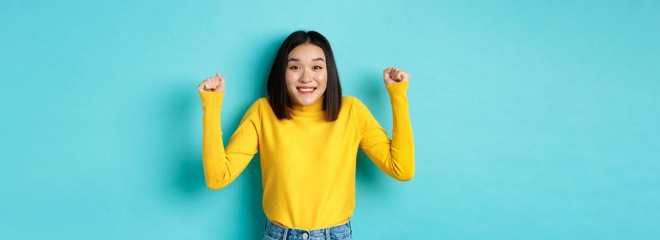 Hopeful asian girl winning prize, clench fists and smiling happy at camera, triumphing of achievement and success, standing over blue background