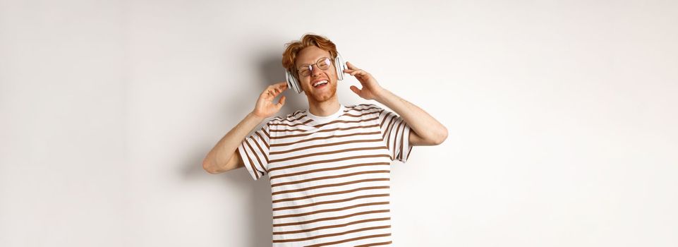 Technology concept. Happy redhead man listening music in headphones and singing along, standing over white background.