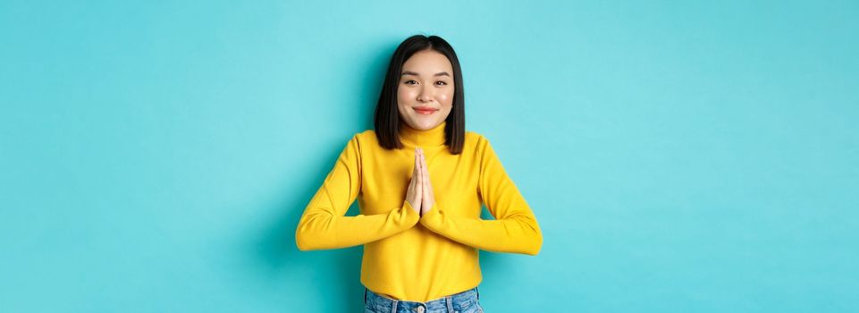 Cute asian woman in trendy outfit saying thank you, holding hands in namaste, begging gesture, smiling grateful at camera, standing over blue background