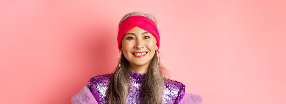 Close-up of trendy asian senior woman with grey hair and stylish disco dress, smiling happy at camera, standing over pink background