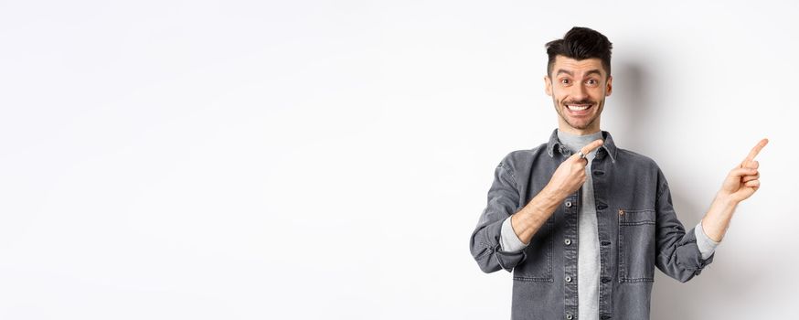 Cheerful stylish man with moustache and white smile, pointing fingers right, inviting to look here, standing on white background