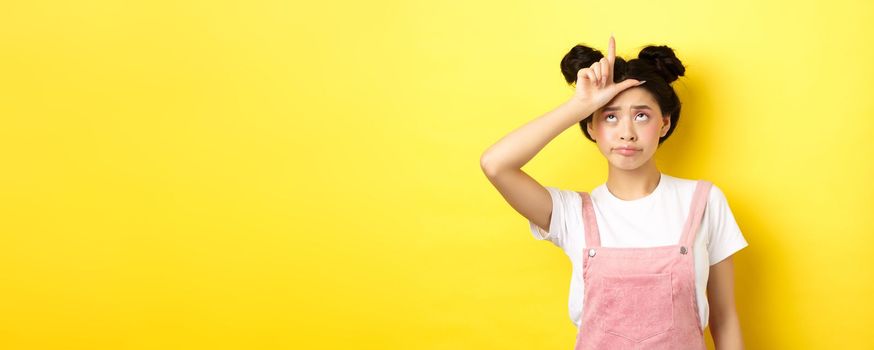 Sad girl showing loser sign on forehead and sulking upset, feeling disappointed in herself, standing on yellow background