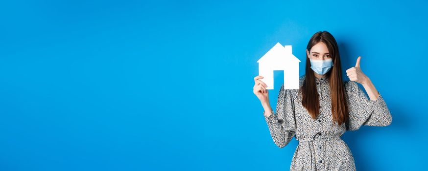 Real estate, covid-19 and pandemic concept. Candid woman in medical mask showing thumb up and paper house cutout, reanting apartment, standing on blue background