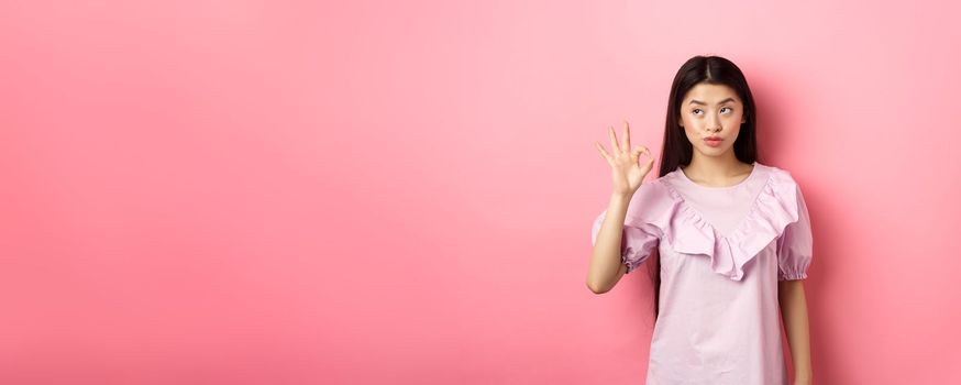 Sassy asian woman in dress show okay sign, looking aside, assure everything good, guarantee all OK, standing pleased on pink background