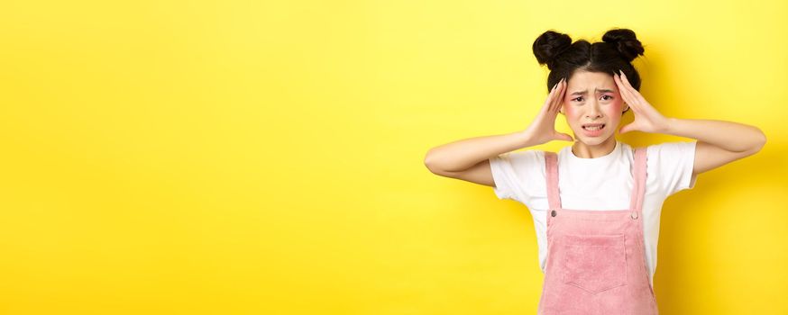 Frustrated asian woman touching head and looking troubled, having problem, standing anxious on yellow background