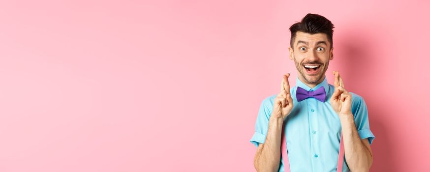 Happy guy making wish, holding fingers crossed for good luck and smiling at camera, waiting for good news, standing over pink background
