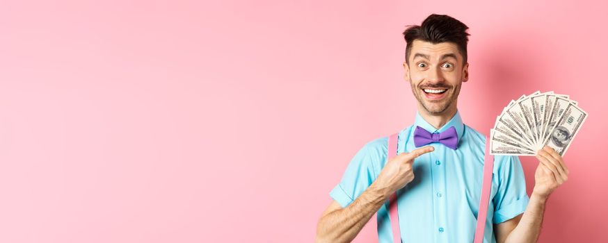 Portrait of cheerful guy with moustache and bow-tie pointing at money, holding dollars and smiling excited, standing over pink background
