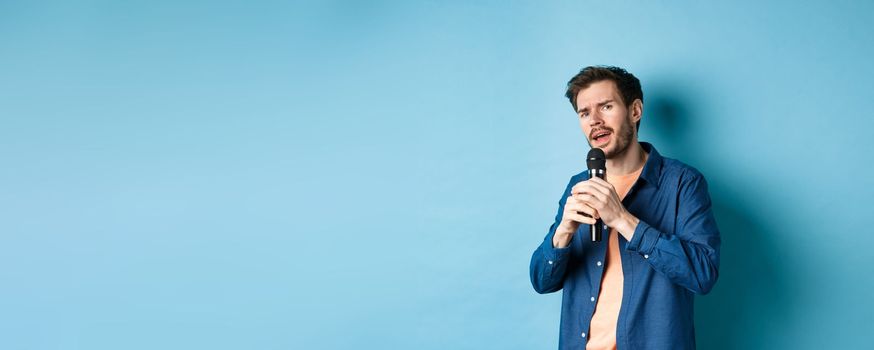 Young man singing song in microphone, playing karaoke, standing on blue background