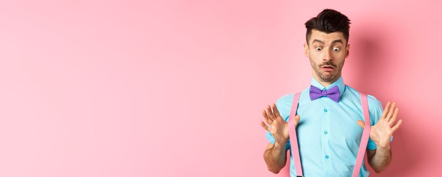 Funny guy with moustache and bow-tie, adjusting his suspenders and looking down with confused and surprised face, standing over pink background