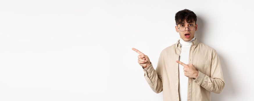 Portrait of young man in glasses drop jaw impressed, pointing fingers left at empty space with amazed face, standing on white background