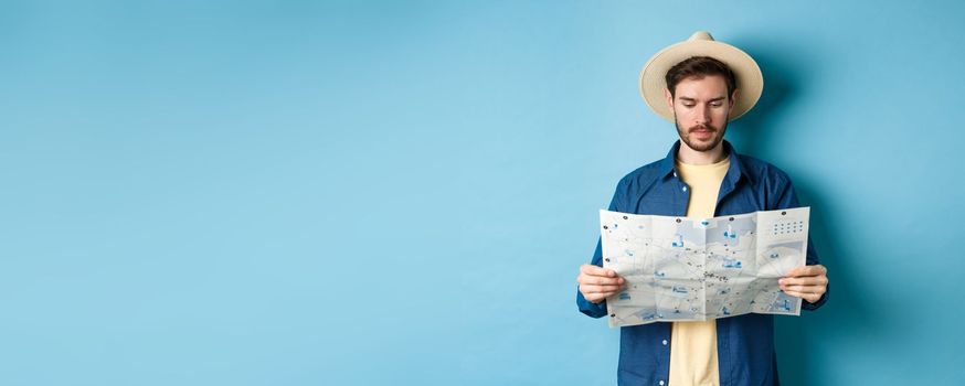 Handsome tourist with summer hat reading map, looking for sightseeing on vacation, standing on blue background