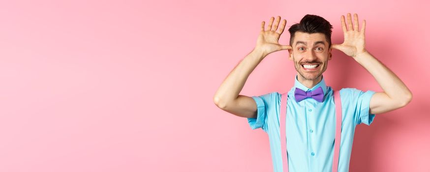 Silly and childish guy in suspenders and bow-tie, mocking someone, fooling around and making funny gestures, standing on pink background