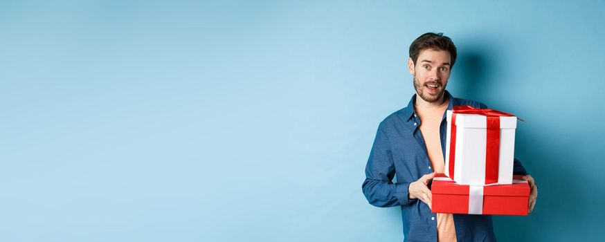 Handsome bearded guy holding Valentines day gifts for lover, standing with presents in boxes and looking at camera, blue background.